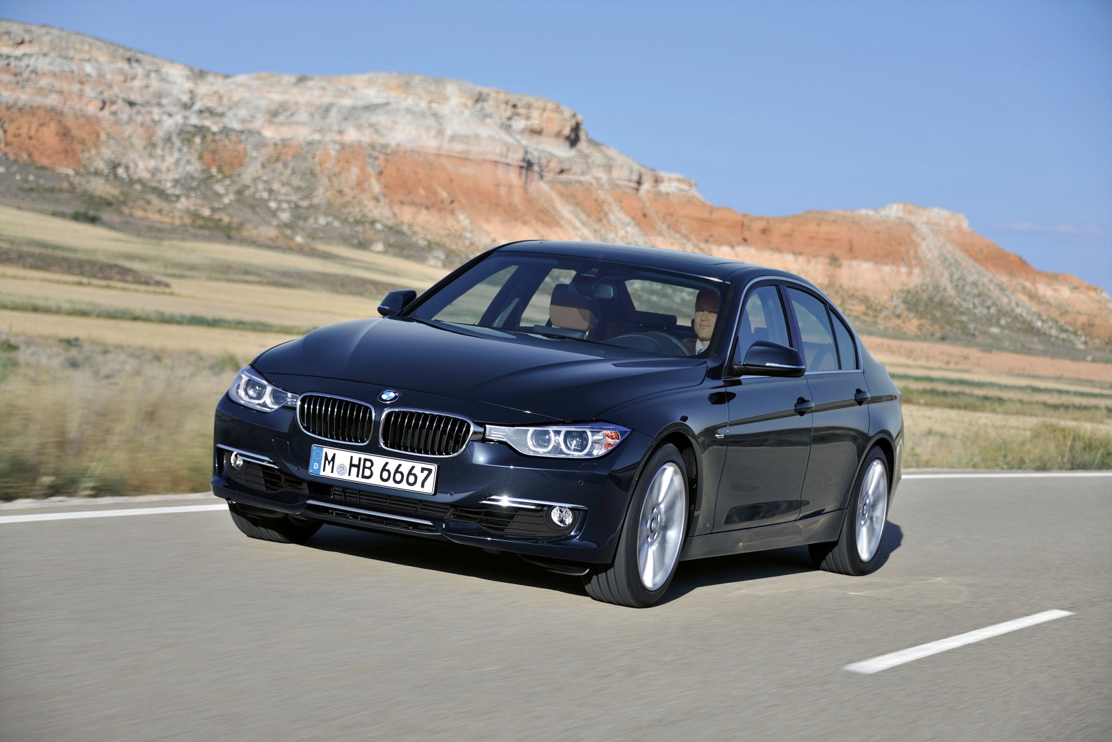 BMW Serie 3 F30 oscuro en carretera, vista delantera de tres cuartos junto a una ladera rocosa